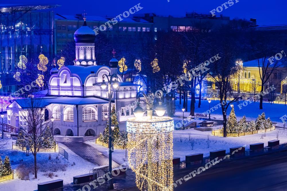 На фотографии изображён православный храм, окружённый снежным парком Зарядье. Вокруг здания установлены праздничные огни и светящиеся конструкции, подчёркивающие зимнее настроение. Освещение фасада храма и украшенные деревья создают атмосферу уюта и праздника. Здание из стекла на заднем плане добавляет современный акцент в традиционный городской пейзаж. Это место прекрасно передаёт гармонию старинной архитектуры и современного дизайна Москвы.
