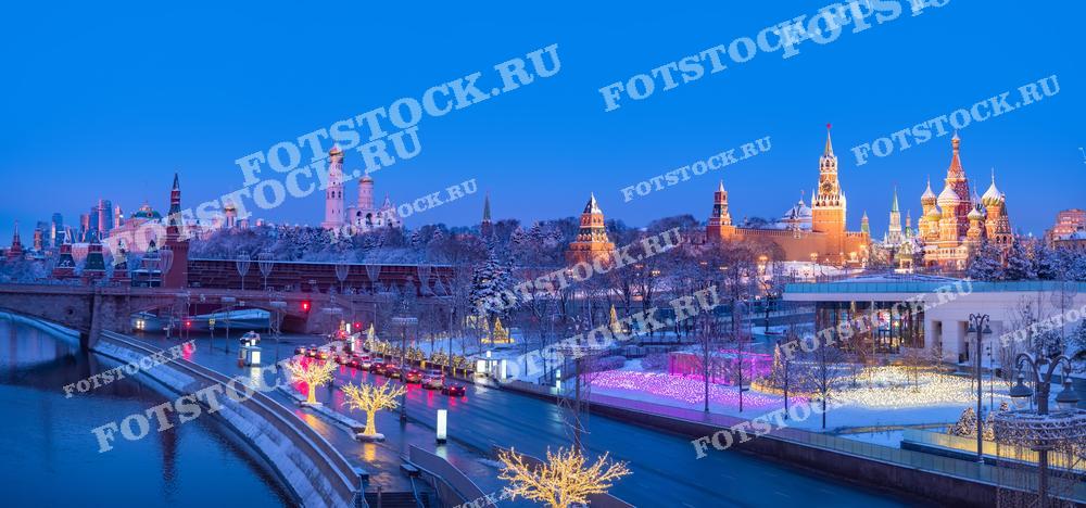 Фотография демонстрирует Москву ранним утром в зимнее время. На заднем плане расположены знаменитые достопримечательности: Кремль с его величественными башнями и куполами Собора Василия Блаженного. Справа — парк Зарядье, украшенный новогодними огнями и световыми инсталляциями. Набережная Москвы-реки вдоль дороги украшена светящимися деревьями, которые подчеркивают зимнюю праздничную атмосферу столицы. Тихий утренний свет придает фотографии мягкость и спокойствие.