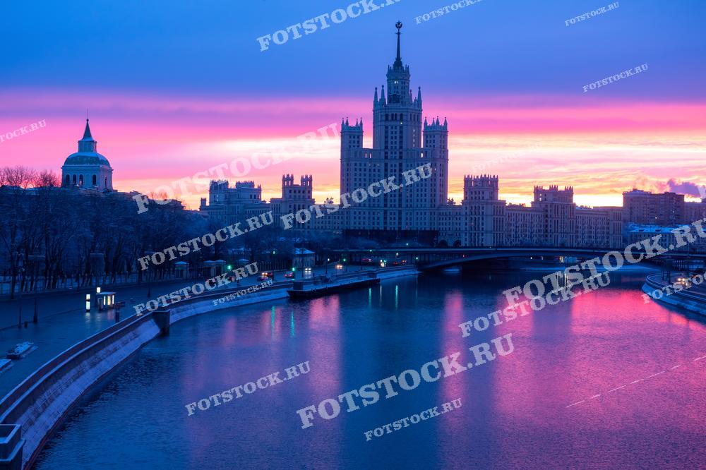 На фотографии изображен один из символов Москвы — сталинская высотка. Ранний зимний рассвет окрашивает небо в яркие розово-фиолетовые оттенки, которые отражаются в водах Москвы-реки. На заднем плане виден мост и здания советской архитектуры, которые подчеркивают исторический облик района. Левее расположено купольное здание с утонченными линиями, дополняющее атмосферу спокойствия и тишины. Этот утренний пейзаж передает красоту зимней Москвы в первые часы дня.
