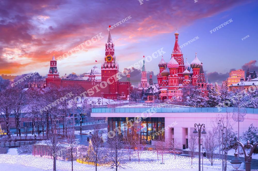 На переднем плане фотографии — Парк Зарядье, украшенный зимней подсветкой и укрытый снегом. В центре кадра возвышаются башни Кремля, включая Спасскую башню с часами. Рядом расположен красочный Собор Василия Блаженного, один из символов Москвы. На фоне неба заката розовые и сиреневые оттенки создают сказочную атмосферу. Видна часть Красной площади и древних стен Кремля. Зимний пейзаж подчёркивает величие архитектуры и праздничное настроение города.