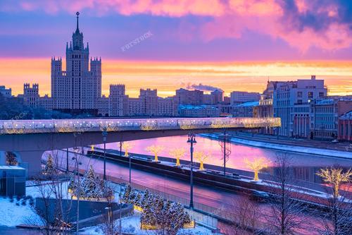 На фотографии запечатлен уникальный городской пейзаж Москвы с плавающим мостом и сталинской высоткой на фоне заката. 
Эта фотография идеально подходит для использования в туристических проспектах, рекламе городских мероприятий, а также для создания атмосферного фона в изданиях, посвященных Москве. Она может быть использована как иллюстрация к статьям о городской архитектуре и туризме.