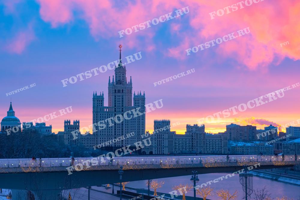 На фотографии изображен восход над одним из высотных зданий Москвы — знаменитой сталинской высоткой. Зимнее утро придаёт городу мягкий, розовый оттенок, подчеркивая архитектурную красоту и величие зданий сталинского ампира. Река под заснеженным мостом и приглушённое освещение добавляют картине умиротворённость. Эта фотография может быть использована в туристических каталогах, архитектурных обзорах и культурных публикациях, акцентирующих на историческом наследии Москвы.
