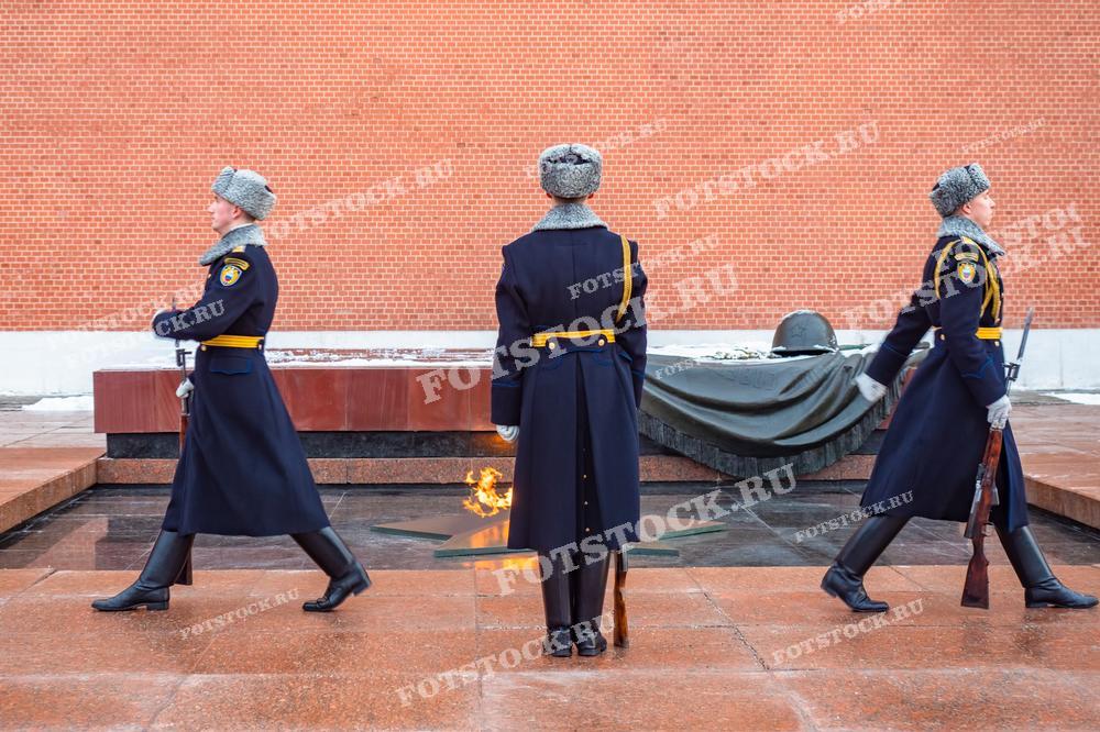 На фотографии изображена церемония смены караула у Вечного огня в Александровском саду, расположенном у стен Московского Кремля. Три гвардейца в зимней форме шагом проходят мимо мемориала, где горит Вечный огонь в честь павших солдат Великой Отечественной войны.