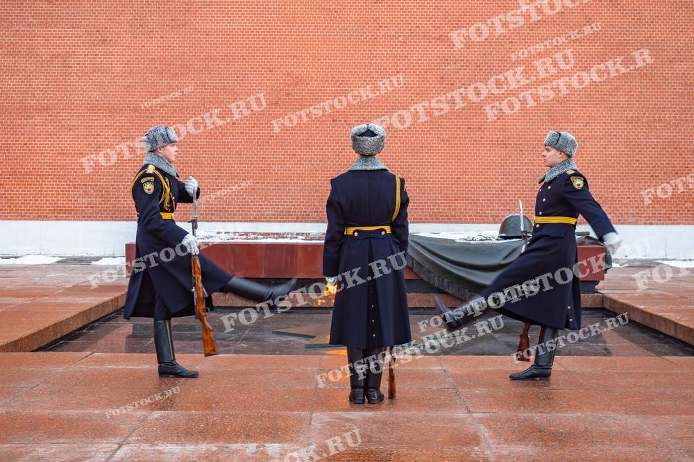 На фотографии запечатлен момент смены караула у Вечного огня в Александровском саду. Три гвардейца в зимней форме выполняют торжественные движения перед мемориалом. Вечный огонь, символизирующий память о павших воинах, горит на переднем плане.