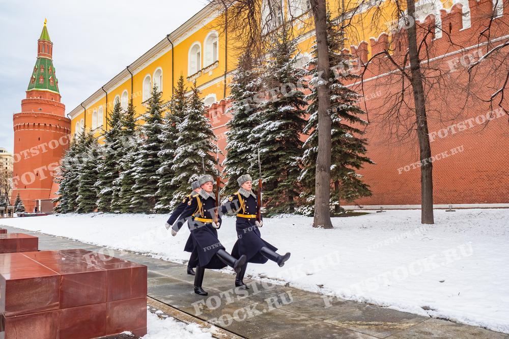 На фотографии запечатлен момент смены караула у Вечного огня в Александровском саду. Три гвардейца в зимней форме выполняют торжественные движения перед мемориалом. Вечный огонь, символизирующий память о павших воинах, горит на переднем плане.