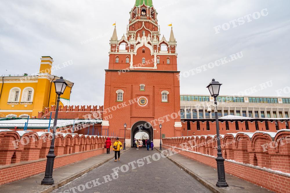На фотографии изображены ворота, ведущие в Московский Кремль. Ворота украшают красные кирпичные стены с часами и зубчатым верхом. Пешеходы проходят через ворота, добавляя жизни и движения в кадр.