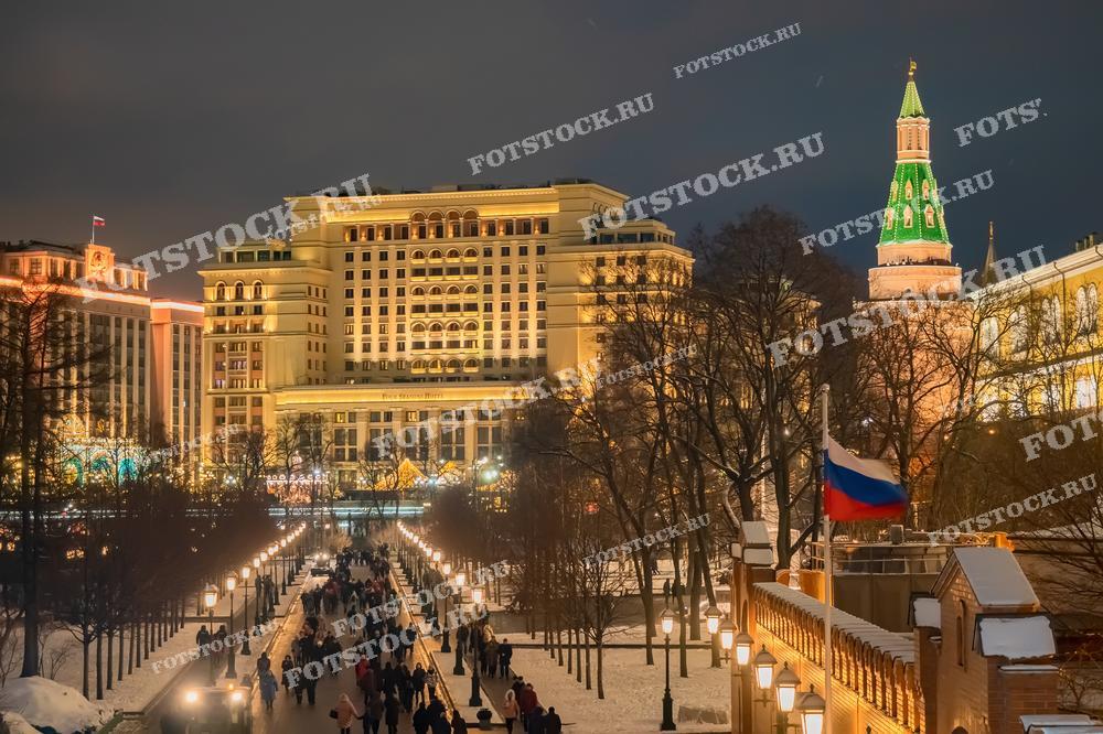 На фотографии изображен ночной вид на центр Москвы зимой. В кадре видна одна из башен Кремля с ярко-зеленым шпилем. Освещенные здания и улицы создают праздничную атмосферу. Пешеходы прогуливаются по Александровскому саду, наслаждаясь видами и зимним воздухом. Флаги России развеваются на фоне освещенных зданий, подчеркивая торжественность момента. Зимняя ночь наполняет снимок уютом и теплом огней.
