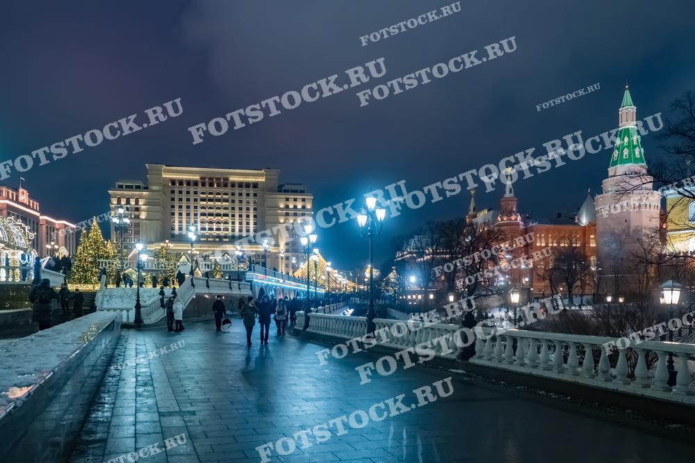 На фотографии изображена Манежная площадь зимой в вечернее время. В центре кадра возвышается отель "Москва", украшенный праздничной иллюминацией. Справа виднеются Кремлевские башни с зелеными шпилями. Уличные фонари и гирлянды освещают площадь, создавая теплую и уютную атмосферу. Пешеходы прогуливаются по заснеженным дорожкам, наслаждаясь видом на праздничную Москву и вечерние огни.