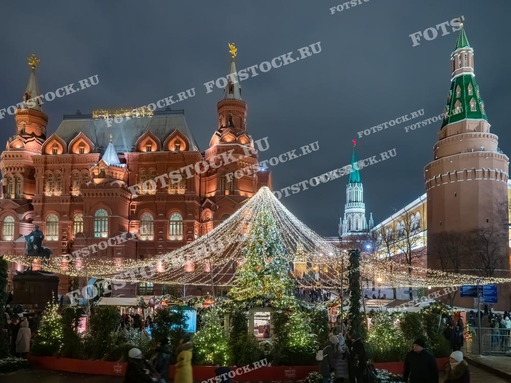 На фотографии изображена праздничная ночь на Красной площади в Москве. Центр кадра украшен новогодней иллюминацией с гирляндами и праздничными огнями. Величественные здания Государственного исторического музея и Кремлевских башен создают атмосферу исторической значимости. Зеленые шпили и красные стены Кремля подсвечены, добавляя яркости и торжественности. Пешеходы гуляют по площади, наслаждаясь новогодними украшениями и праздничной атмосферой зимней Москвы.