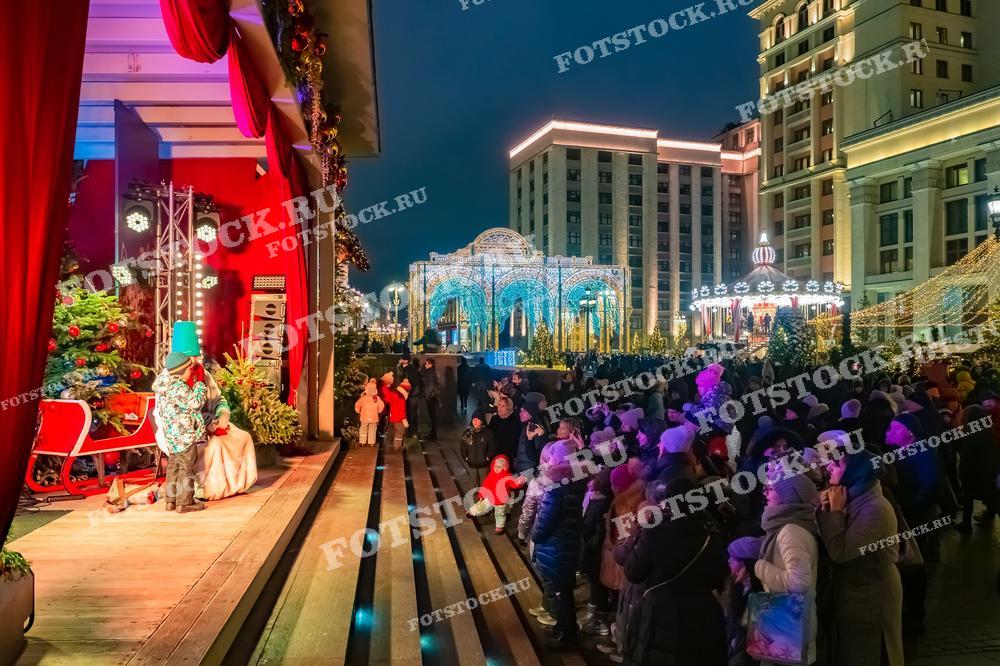 На фотографии изображен новогодний концерт на Манежной площади в Москве. Праздничная сцена украшена гирляндами, елками и красными занавесками. На сцене выступают артисты в новогодних костюмах, развлекая собравшихся зрителей. Площадь освещена праздничными огнями и иллюминацией, создавая волшебную атмосферу. Вдалеке видны украшенные здания и арки, добавляющие еще больше праздничного настроения. Люди наслаждаются концертом и праздничным вечером в центре Москвы.