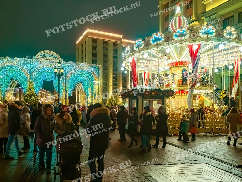 На фотографии изображены новогодние гуляния на Манежной площади в Москве. В центре кадра видна яркая карусель, украшенная огнями и праздничными декорациями. Справа виднеются иллюминированные арки, создающие волшебную атмосферу. Люди прогуливаются по площади, наслаждаясь зимним вечером и праздничным настроением.