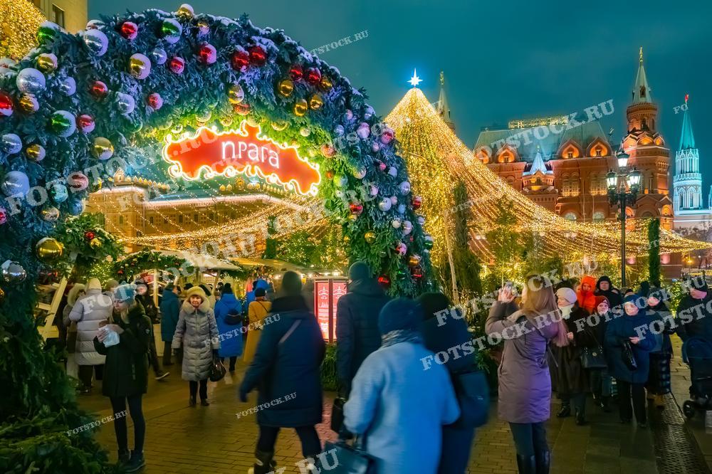 На фотографии изображена новогодняя ярмарка на Манежной площади в Москве. Центральное место занимает украшенная гирляндами арка, приветствующая гостей. Яркая новогодняя иллюминация создает волшебную атмосферу праздника. Люди гуляют по ярмарке, наслаждаясь зимним вечером и праздничными угощениями.