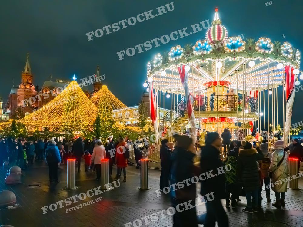 На фотографии изображена новогодняя ярмарка на Манежной площади в Москве. В центре кадра - ярко освещенная карусель, украшенная праздничными декорациями. Вокруг неё прогуливаются люди, наслаждаясь атмосферой праздника. Позади карусели виднеется большая новогодняя елка, украшенная гирляндами и огнями, а также здания Государственного исторического музея и Кремлевских башен. Праздничная иллюминация и зимний вечер создают волшебное настроение.