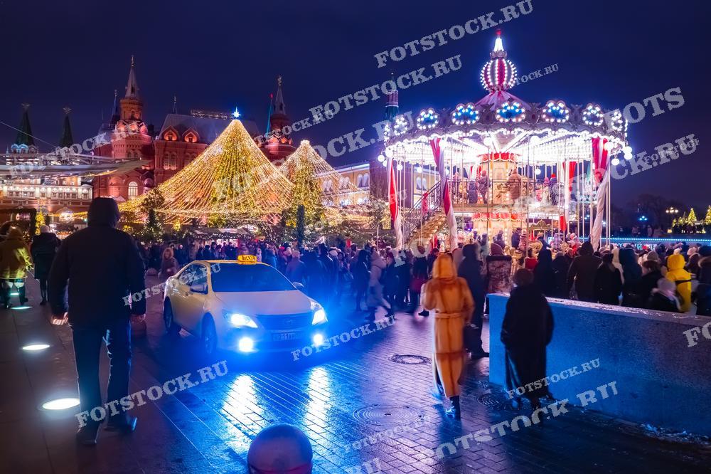 На фотографии изображена праздничная карусель и новогодние огни на Манежной площади в Москве. Карусель украшена гирляндами и огнями, создавая волшебную атмосферу праздника. Вокруг неё собралось много людей, наслаждающихся зимним вечером. Позади видна большая новогодняя елка, украшенная огнями, а также здания Государственного исторического музея и Кремлевских башен.