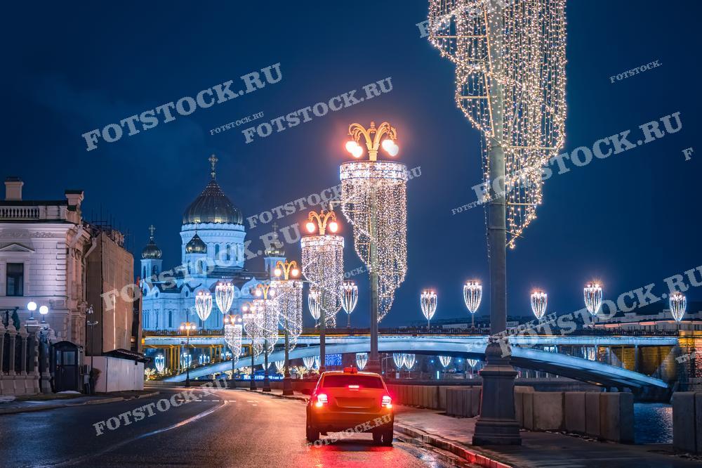 На фотографии изображена новогодняя иллюминация на набережной Москвы-реки с видом на Храм Христа Спасителя. Светящиеся украшения и огни города создают праздничную атмосферу зимней Москвы. Фотография подходит для использования в материалах о новогодних праздниках, туристических буклетах и статьях о зимней Москве.