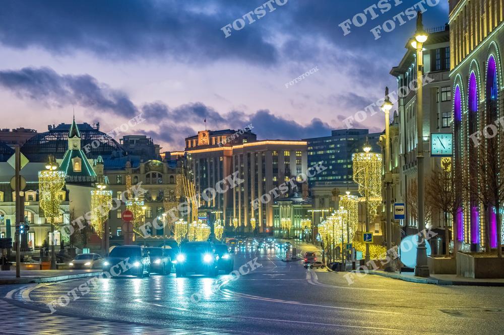 На фотографии изображён вечерний вид одной из центральных улиц Москвы, украшенной новогодней иллюминацией. Светящиеся деревья вдоль дороги придают улицам праздничное настроение. Архитектурные элементы зданий гармонично сочетаются с современными украшениями. На заднем плане можно увидеть переливающееся закатное небо, которое подчёркивает атмосферу зимнего вечера. Этот уголок Москвы отлично передаёт сочетание городской суеты и спокойного праздничного настроения.