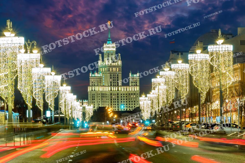 На фотографии изображена московская улица, украшенная новогодней иллюминацией. Вдоль улицы горят праздничные огни, создавая атмосферу праздника. На заднем плане видна одна из сталинских высоток, освещенная в ночное время.