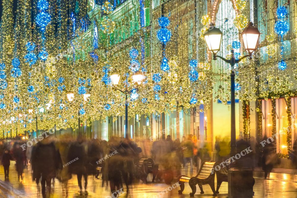 На фотографии изображена улица в центре Москвы в период новогодних праздников. Улица сияет яркими огнями: гирлянды и световые шары украшают пространство над головами прохожих. Это одно из самых популярных мест для прогулок и фотосессий в зимнюю столицу. По обеим сторонам улицы расположены исторические здания и витрины магазинов, оформленные в праздничном стиле. Видно, как множество людей неспешно гуляют, наслаждаясь атмосферой зимнего торжества. Освещённые фонари и мягкий свет создают ощущение уюта и праздника.