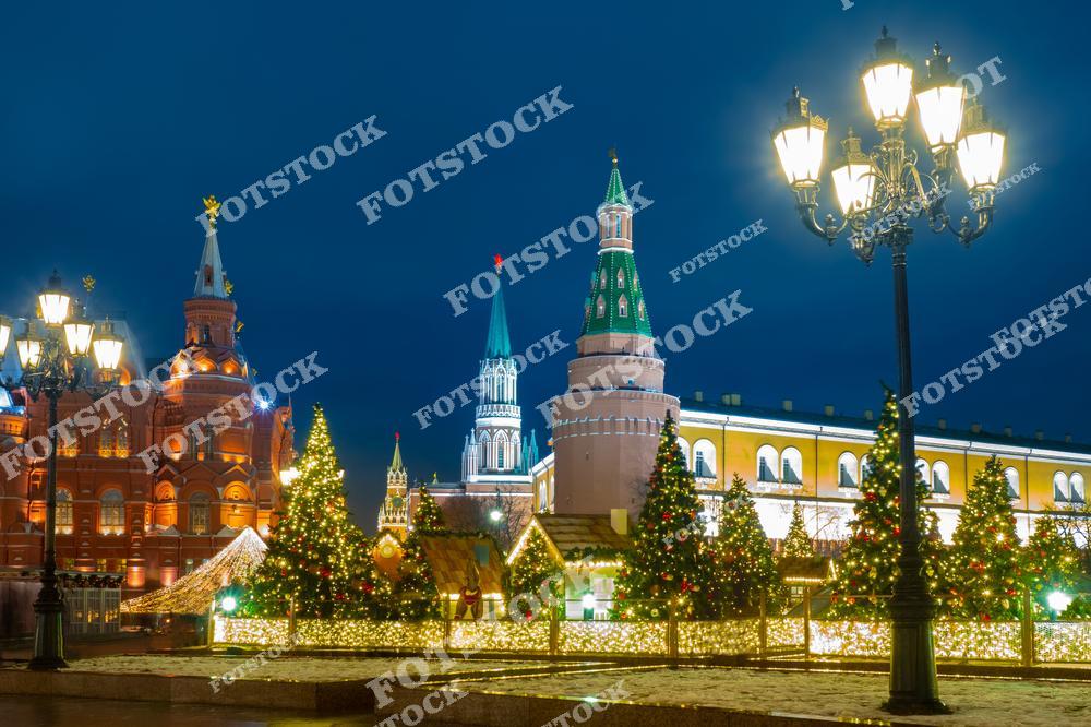 На фотографии изображён Московский Кремль в новогоднюю ночь, когда его башни подсвечены огнями. Вдоль площади стоят ёлки, украшенные яркими гирляндами, создающими атмосферу праздника. Это зимний вечер, когда праздничное освещение подчеркивает величие Кремля и делает Москву особенно привлекательной. Декорации, фонари и украшения на площади добавляют сказочности и чарующего света.