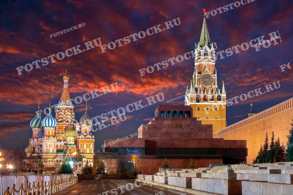 На фотографии изображен вечерний вид на Красную площадь в Москве во время заката. В центре кадра видны Собор Василия Блаженного и Спасская башня Кремля, освещенные закатными лучами. На переднем плане находится Мавзолей Ленина.