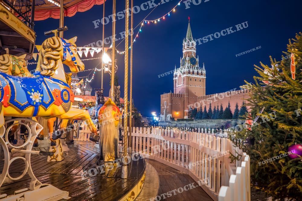 На фотографии изображена новогодняя карусель на Красной площади в Москве. На заднем плане видна Спасская башня Кремля. Светящиеся гирлянды и праздничные украшения создают атмосферу новогоднего праздника. Фотография подходит для использования в материалах о новогодних праздниках, туристических буклетах и статьях о зимней Москве.