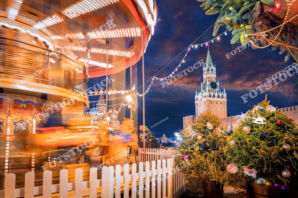 На фотографии изображена Красная площадь в Москве в новогоднюю ночь. В центре композиции — карусель, на которой создаётся эффект движения, благодаря чему в кадре появляется динамичность и праздничная атмосфера. Вокруг карусели стоят ёлки, украшенные гирляндами и новогодними игрушками. На фоне видна Спасская башня Кремля, которая также подсвечена огнями, создавая волшебную атмосферу праздника. Яркие огоньки и украшения дополняют зимний пейзаж.