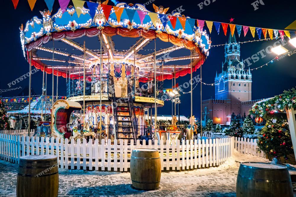 На фотографии изображена новогодняя карусель на Красной площади в Москве, украшенная яркими огоньками и флажками. Вокруг карусели стоят ёлки, украшенные гирляндами и новогодними игрушками, создающими атмосферу праздника. На заднем плане видна Спасская башня Кремля, которая также подсвечена огнями, добавляя волшебства в эту зимнюю ночь. Оформление площади и рождественская атмосфера делают эту сцену яркой и полной жизни.