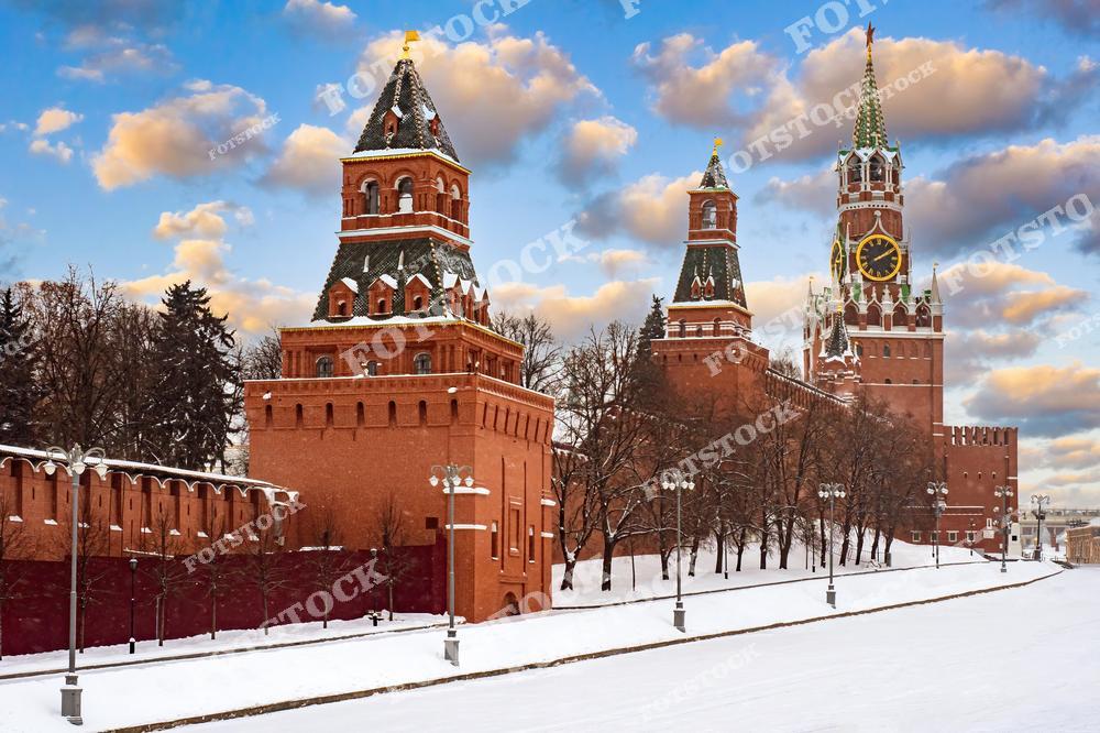 На фотографии изображён Московский Кремль, главный символ российской государственности. В кадре отчётливо видна Спасская башня с курантами — одна из самых узнаваемых достопримечательностей Москвы. Рядом расположены другие кремлёвские башни, окружённые краснокирпичной крепостной стеной. Земля покрыта чистым белым снегом, а небо украшено живописными облаками, подчёркивающими торжественность зимнего пейзажа. Это место входит в список Всемирного наследия ЮНЕСКО и является обязательным для посещения туристами. Зимой Кремль приобретает особое очарование, подчеркивающее его величие и историческое значение.
