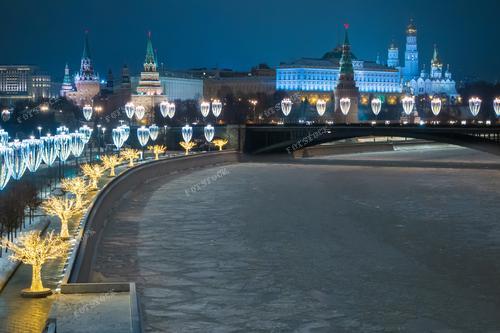 На фотографии представлен зимний пейзаж Москвы: замерзшая река Москва, мерцающая новогодняя иллюминация и Кремль, утопающий в снегу. Снимок передает спокойствие и красоту зимнего вечера в российской столице. Он идеально подходит для использования в туристических брошюрах, новогодних открытках, а также в рекламе зимнего отдыха в Москве. Фотография станет прекрасным дополнением к материалам о российских городах и зимних праздниках.