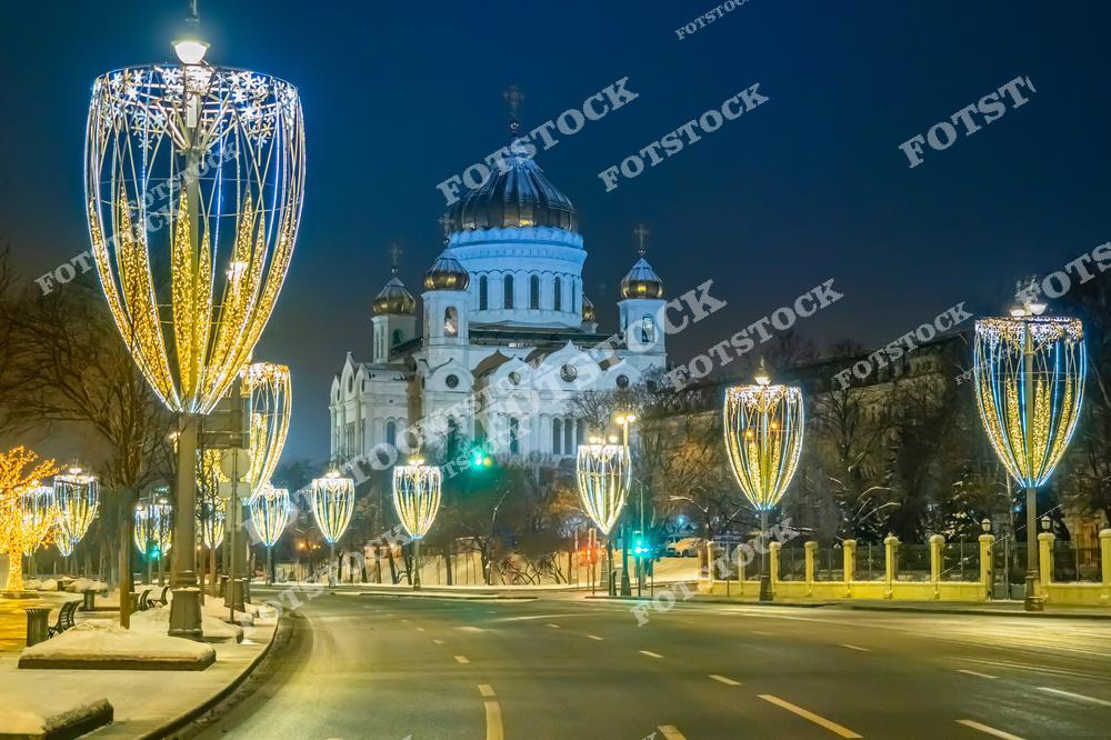 На фотографии запечатлён Храм Христа Спасителя, один из главных символов Москвы, украшенный вечерней подсветкой. Вдоль дороги установлены световые конструкции в форме изящных гирлянд, создающих праздничное настроение. Снежный покров подчёркивает зимнюю атмосферу, а пустынная улица добавляет ощущение спокойствия. Архитектура храма гармонирует с современными элементами праздничного оформления столицы. Этот вид передаёт красоту ночной Москвы в период зимних праздников.