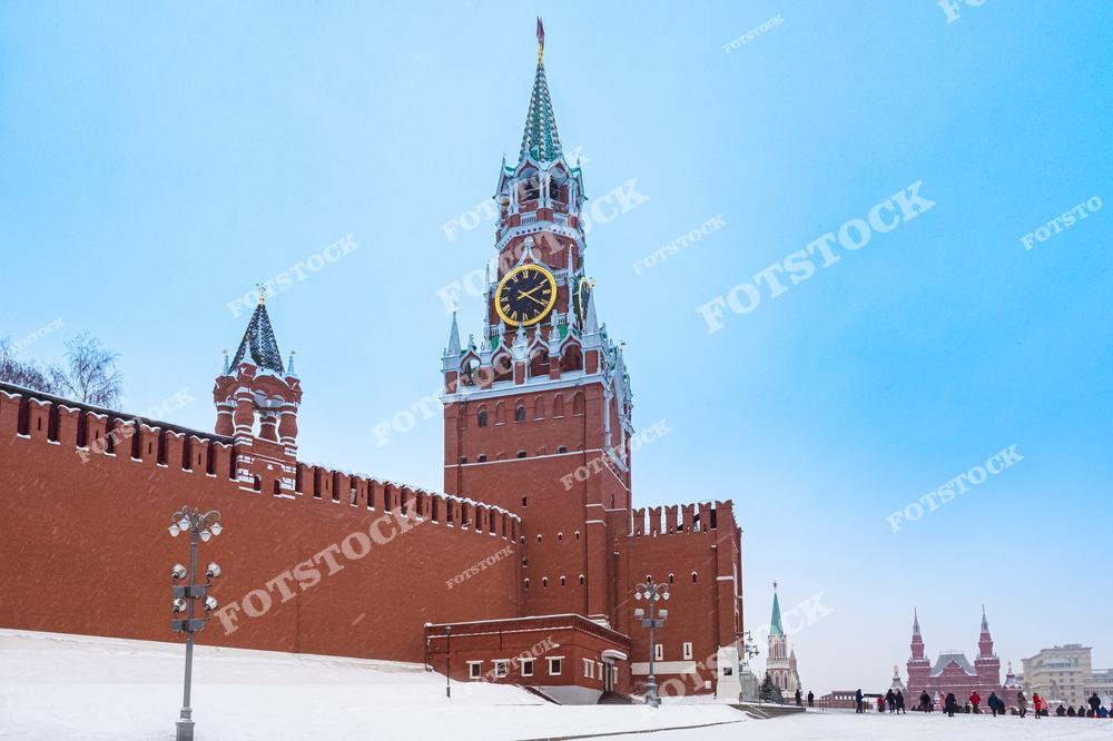 На фотографии изображён Кремль в Москве с видом на Спасскую башню, которая украшена ярким часами на фоне зимнего неба. На переднем плане видна стена Кремля, покрытая снегом, и уличные фонари, создающие контраст с холодным голубым небом. Слева и справа виднеются другие части Кремля, подчеркивающие величие и историческую значимость этого символа столицы России. Снежный пейзаж и ясное небо придают картине чистоту и спокойствие.