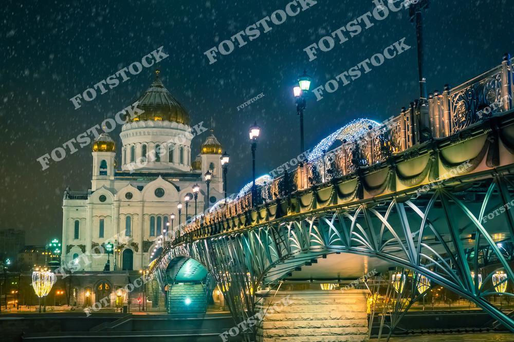 На фотографии изображён Храм Христа Спасителя, освещённый мягким светом в ночное время. Справа проходит пешеходный мост, украшенный гирляндами и световыми элементами, создающими праздничное настроение. Снегопад добавляет атмосферности этой зимней сцене, подчёркивая тишину и величие места. Отражения огней на воде и архитектурные детали моста делают картину живописной.