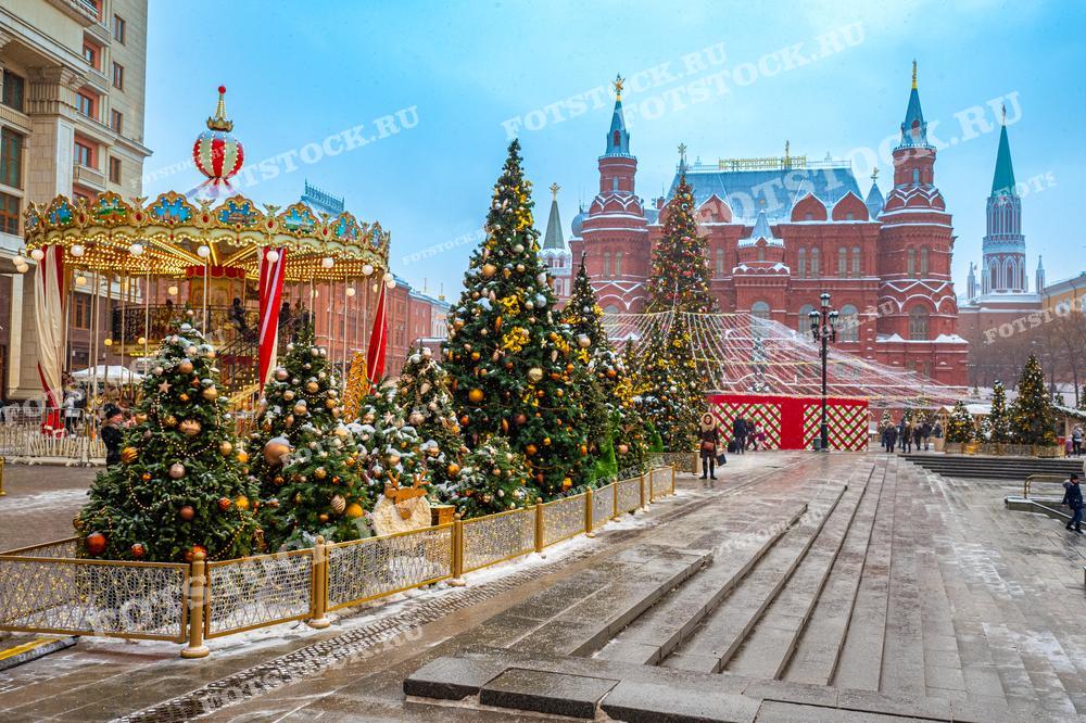 На фотографии изображена новогодняя карусель и праздничные украшения на Манежной площади в Москве. Яркая карусель с красными и золотыми элементами привлекает внимание. Вокруг установлены украшенные елки с гирляндами и игрушками, создавая волшебную атмосферу. На заднем плане виднеется Государственный исторический музей с его красными стенами и башнями. Справа можно увидеть Кремлевские башни с зелеными шпилями. Люди прогуливаются по площади, наслаждаясь зимним днем и праздничным убранством города.