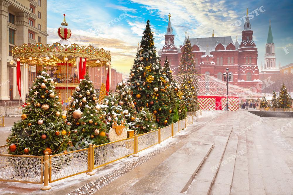 На фотографии изображена празднично украшенная площадь в Москве. На переднем плане расположены ёлки, украшенные яркими шарами и гирляндами, создающими новогоднюю атмосферу. Слева видна карусель, украшенная красными и золотыми элементами, а на заднем плане можно увидеть здание Государственного исторического музея и Кремль. Лёгкий снежный покров на земле подчёркивает зимний пейзаж, а мягкий свет заката добавляет тёплые оттенки в праздничную сцену.