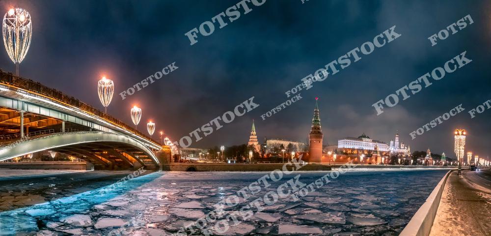На фотографии запечатлена Москва-река, покрытая ледяными дрейфующими льдинами, на фоне величественного Кремля. Слева расположен подсвеченный мост с декоративными светильниками, добавляющими праздничный акцент. Башни Кремля и подсветка зданий на набережной создают атмосферу зимнего величия Москвы. Тёмное вечернее небо подчёркивает яркость огней и контрастность зимнего пейзажа. Это место является символом исторического и культурного центра столицы.