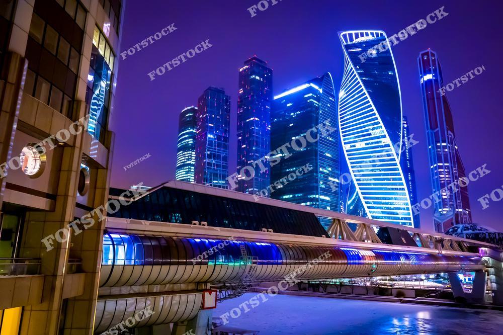 На фотографии изображён деловой центр Москва-Сити в ночном освещении. Высотные здания с уникальной архитектурой, включая башню «Эволюция» с её закрученной формой, ярко подсвечены неоновыми огнями. На переднем плане — футуристический пешеходный мост «Багратион», соединяющий разные части комплекса. Фиолетовый оттенок неба подчёркивает современность и динамичность московского мегаполиса. Это место символизирует деловую активность и прогресс столицы.
