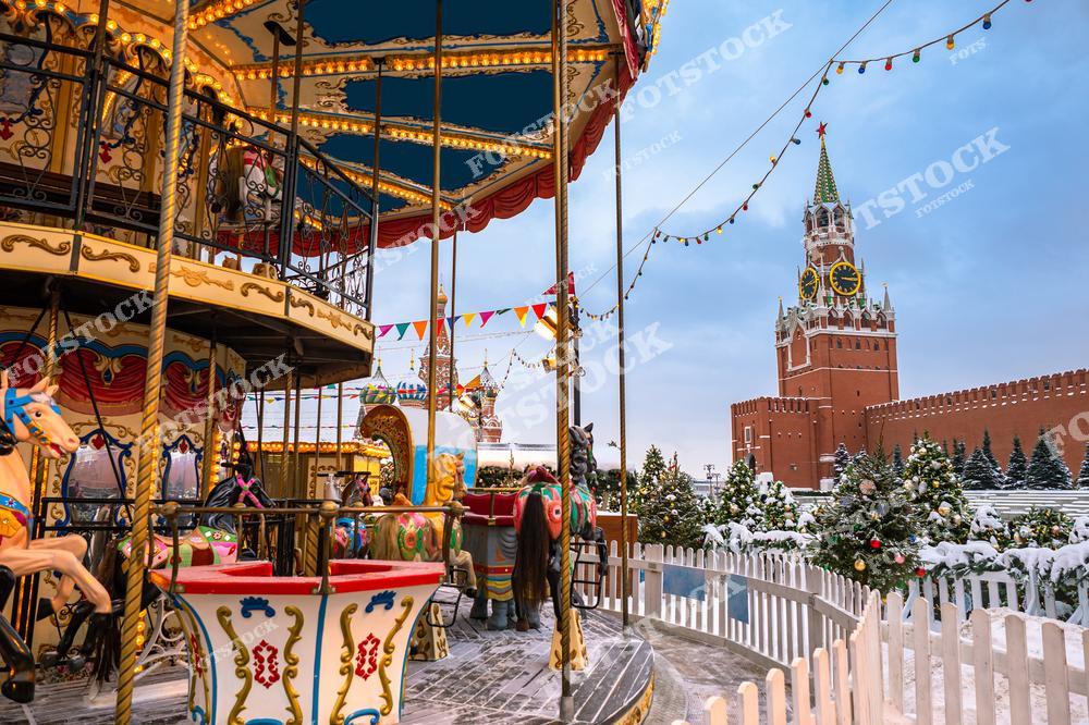 На фотографии изображена праздничная карусель на Красной площади, украшенная яркими гирляндами и лошадками, создающими атмосферу детства и радости. На заднем плане видна Спасская башня Кремля с курантами, возвышающаяся над заснеженным пейзажем. Украшенные ёлки и разноцветные флажки добавляют новогоднего настроения. Светлый зимний день подчёркивает праздничное убранство столицы, превращая это место в центр семейного отдыха и ярмарочной суеты.
