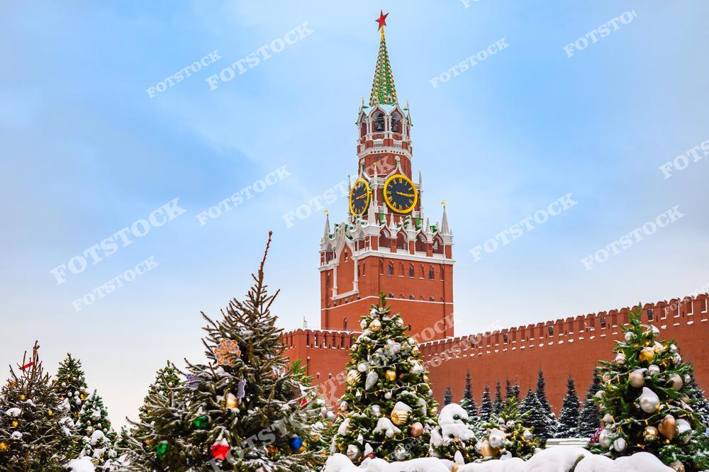 На фотографии изображена Спасская башня Кремля, символ российской столицы. На переднем плане расположены нарядно украшенные новогодние ёлки с гирляндами и золотыми шарами. Красный кирпич кремлёвской стены и знаменитые куранты подчёркивают историческое величие этого места. Снежные ветви и белое зимнее небо создают атмосферу уюта и праздника. Красная площадь превращается в зимнюю сказку, привлекая москвичей и гостей столицы.