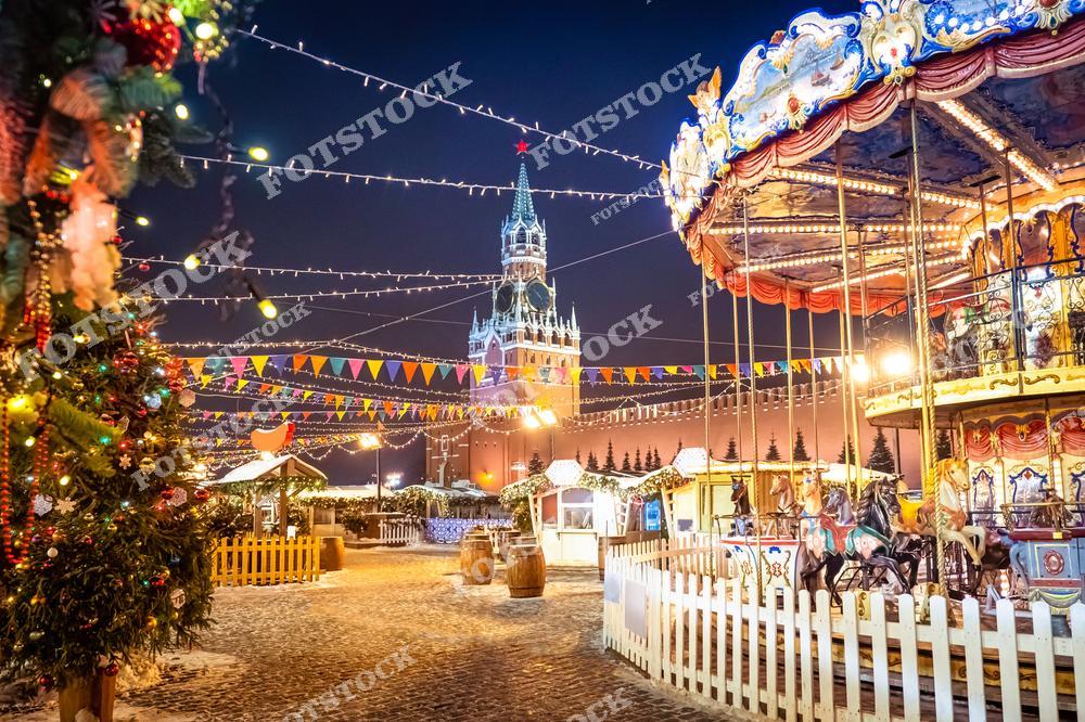 На фотографии изображена Красная площадь в Москве, украшенная к Новому году. На переднем плане видна праздничная карусель с лошадками, ярко освещённая гирляндами и декорациями. Слева расположена украшенная ёлка с гирляндами и шарами. На заднем плане возвышается Спасская башня Кремля с яркой звездой на шпиле. Множество световых гирлянд и украшений создают волшебную атмосферу зимнего праздника.