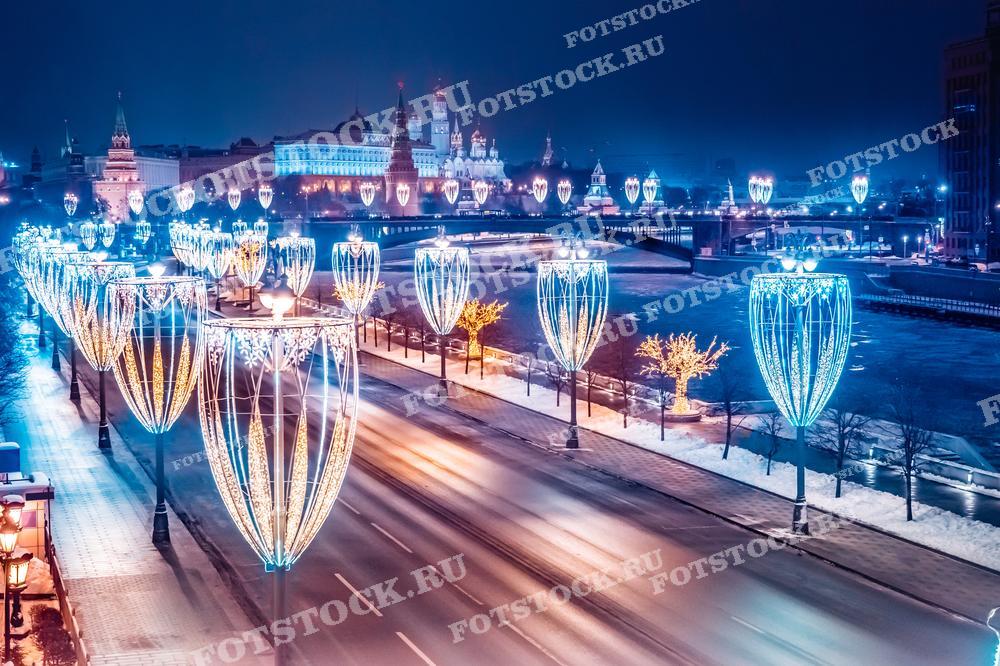На фотографии изображён зимний вечер на одной из набережных Москвы. Над дорогой установлены декоративные светильники в форме фонарей, которые освещают улицу и придают праздничное настроение. На фоне видны Кремль и замерзшая река Москва, что создаёт атмосферу холодной, но великолепной московской зимы. Фотография идеально подойдёт для иллюстрации тем о городской архитектуре или праздничном освещении в статьях о зимних праздниках.