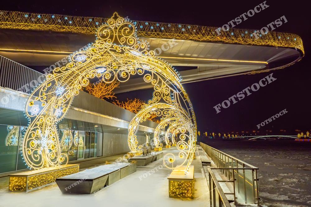 На фотографии изображён парящий мост в парке Зарядье, который украшен световыми арками к новогодним праздникам. Золотистые узоры на арках создают праздничную и сказочную атмосферу. Световые инсталляции подчёркивают современный архитектурный стиль моста, а зимний вечер добавляет картине уюта. Украшенный парк и световые огни делают это место привлекательным для прогулок.