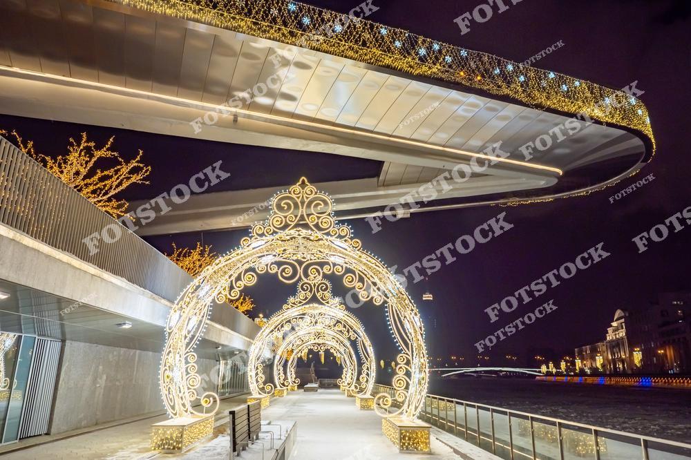 На фотографии запечатлён знаменитый парящий мост в парке Зарядье, украшенный праздничной иллюминацией. На переднем плане расположены сверкающие арки с декоративными узорами, подчёркивающие зимнюю атмосферу. Мост словно парит над набережной, создавая эффект лёгкости и современности. Вдали видны огни города и Москва-реки, добавляющие динамики городскому пейзажу. Это место стало одним из самых популярных для прогулок в зимние праздники.