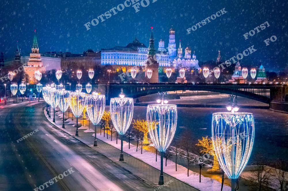 На фотографии изображён ночной вид на Кремль в Москве, украшенный яркой праздничной иллюминацией. Вдоль набережной Москва-реки расположены высокие светящиеся конструкции в форме новогодних украшений, которые создают сказочную атмосферу. Виден заснеженный мост через реку, а падающий снег усиливает ощущение зимней сказки. Кремль сияет в ночной подсветке, подчёркивая его величественную архитектуру. Эта сцена передаёт красоту и уют зимней Москвы во время праздников.