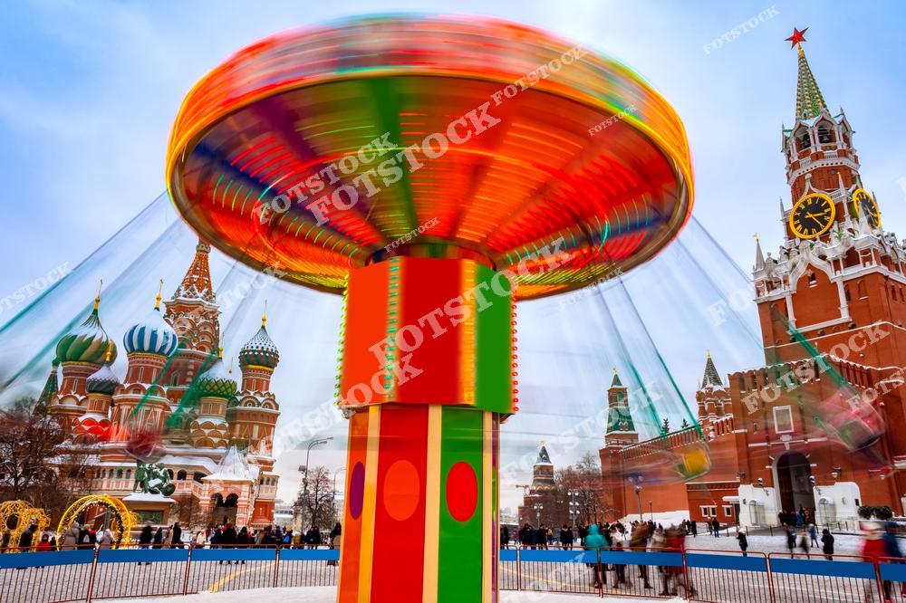 На фотографии изображена яркая карусель на Красной площади, создающая динамичный эффект вращения. На заднем плане видны величественная Спасская башня Кремля и знаменитый Собор Василия Блаженного. Площадь украшена праздничными арками и иллюминацией.