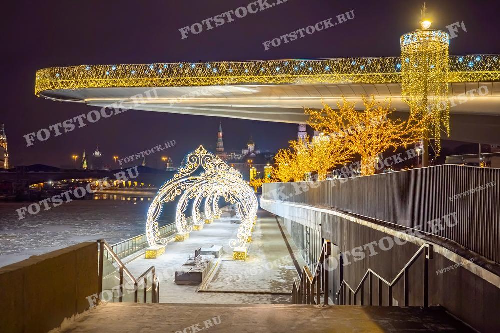 На фотографии изображен зимний вечер в Парке Зарядье, украшенном праздничной иллюминацией. Светящийся мост и декоративные арки создают волшебную атмосферу. Вдалеке видны башни Кремля, отражающие историческое наследие Москвы.