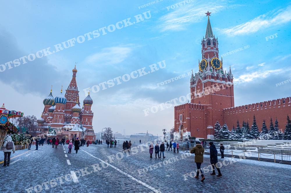 На фотографии изображен зимний день на Красной площади в Москве. Справа расположена Спасская башня Кремля с часами и красной звездой на шпиле. Слева виден Собор Василия Блаженного с его разноцветными куполами. Люди прогуливаются по площади, наслаждаясь зимним днем и архитектурными достопримечательностями. Снежное покрытие и голубое небо создают атмосферу спокойствия и красоты в историческом центре города.