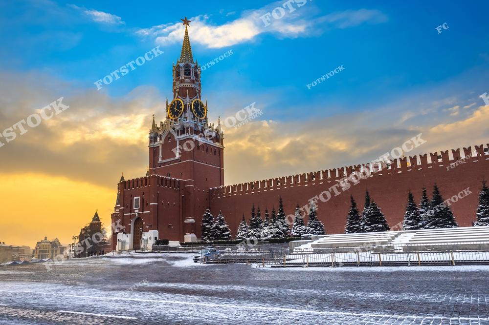 На фотографии изображена Спасская башня Московского Кремля, освещённая мягким светом заката. Заснеженная Красная площадь подчёркивает зимнюю атмосферу. Яркие краски неба — от золотистого до голубого — создают живописный фон, подчёркивающий величие архитектуры. Это спокойный зимний вечер в центре столицы России.