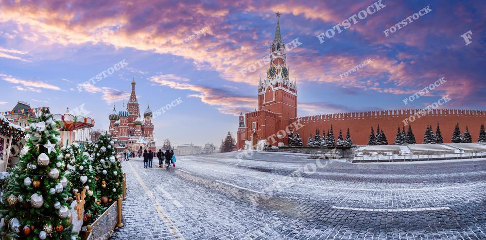 На фотографии изображена Красная площадь в Москве в зимний период, украшенная к праздникам. Слева — наряженные ёлки и часть новогодней ярмарки с каруселью. В центре — Собор Василия Блаженного с его знаменитой архитектурой и яркими куполами. Справа возвышается Спасская башня Кремля с курантами и Кремлёвская стена, украшенная заснеженными ёлками. Мостовая покрыта тонким слоем снега, а небо окрашено в тёплые цвета рассвета. Атмосфера фотографии наполнена тишиной, торжественностью и ощущением зимнего праздника.