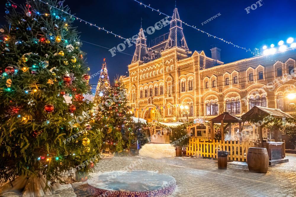 На фотографии изображена празднично украшенная Красная площадь в Москве. На переднем плане видны ёлки, наряженные гирляндами и яркими шарами, создающие атмосферу праздника. На заднем плане — ГУМ, фасад которого украшен иллюминацией, подчёркивающей архитектурные детали здания. В центре композиции расположены новогодние домики и декоративные элементы, дополняющие волшебную картину зимнего вечера. Мягкий свет гирлянд и зимняя атмосфера делают эту сцену уютной и торжественной.