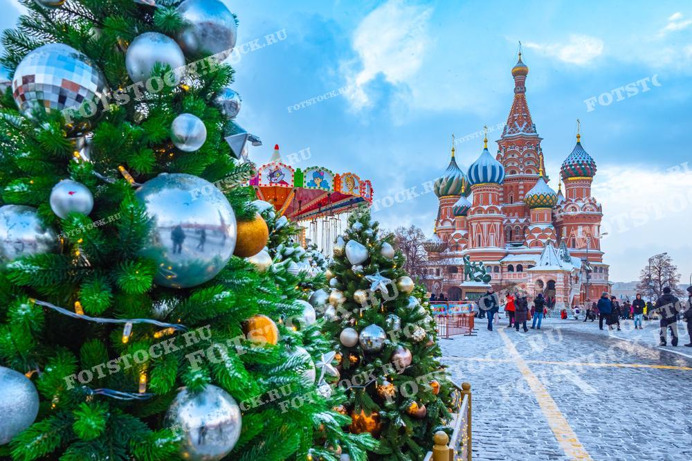 На фотографии изображены новогодние елки на Красной площади в Москве. Елки украшены серебряными шарами и гирляндами, создавая праздничное настроение. На заднем плане виден Собор Василия Блаженного с его яркими куполами. Люди прогуливаются по площади, наслаждаясь зимним днем и праздничными декорациями. Карусель и украшенные деревья добавляют волшебства и радости в атмосферу зимней Москвы.